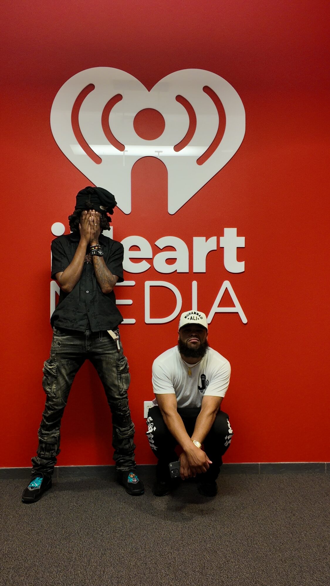 Two people pose in front of a red wall with the iHeartRadio logo and 'iHeartMEDIA' text, one standing and one crouching.