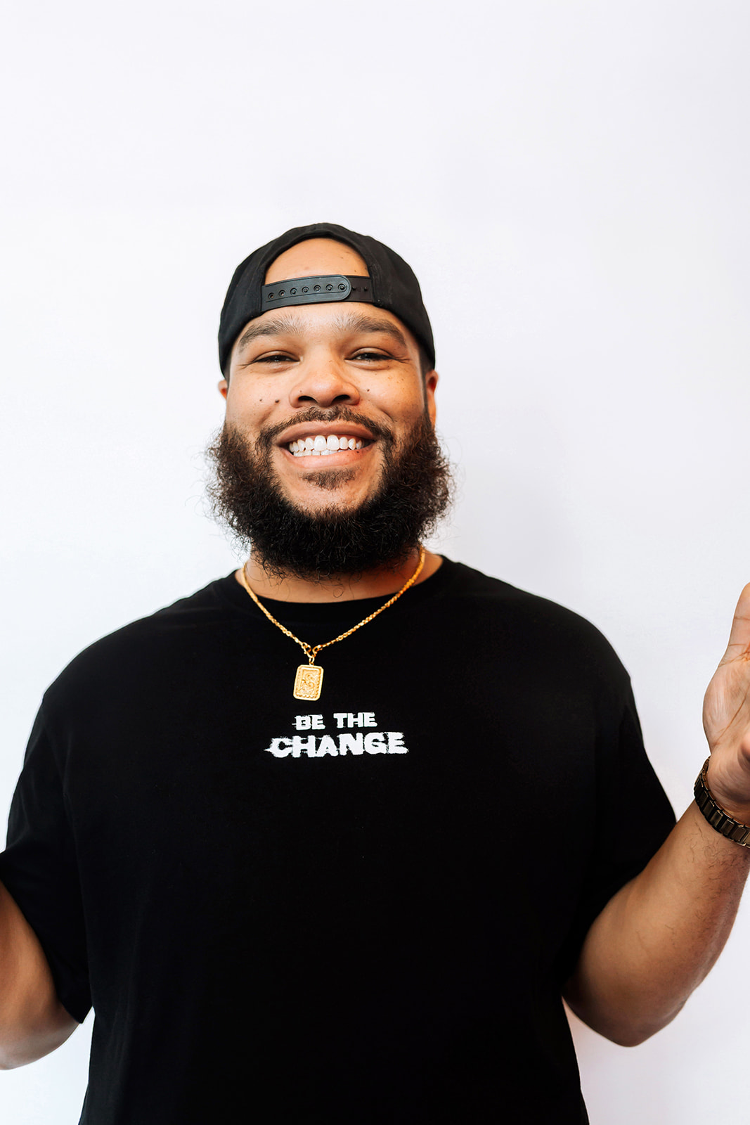 Smiling man with a beard wearing a black cap backward and a black T-shirt with white text, holding a phone.