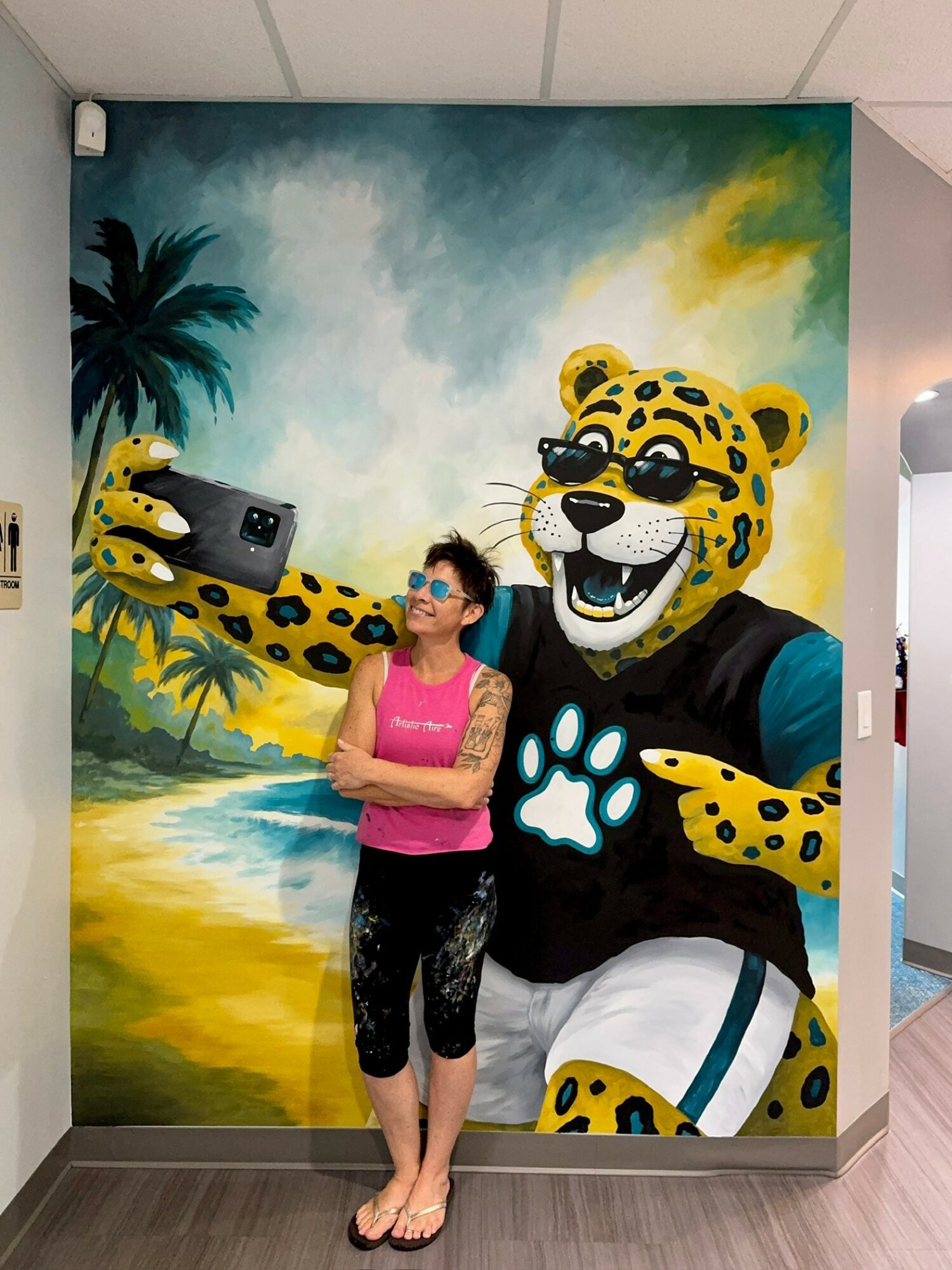 Woman with glasses and pink top stands next to a colorful mural of a smiling leopard wearing sunglasses, in a tropical setting.