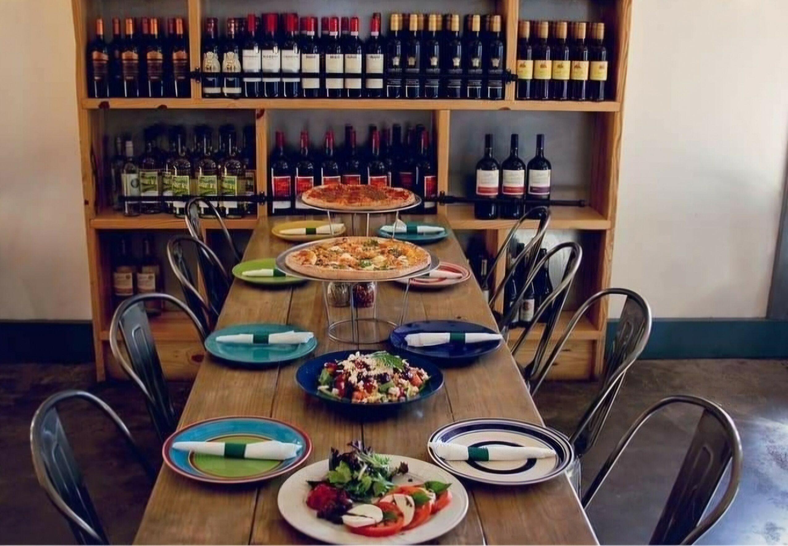Long wooden table set with plates, a pizza, a salad, and a cake, in front of a wine shelf.