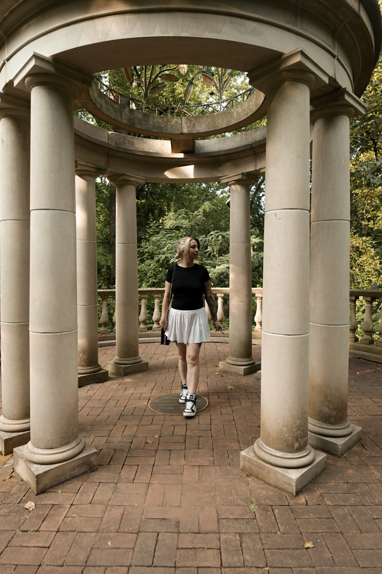 Woman walking through a circular stone pavilion with columns in a park, surrounded by trees and greenery.