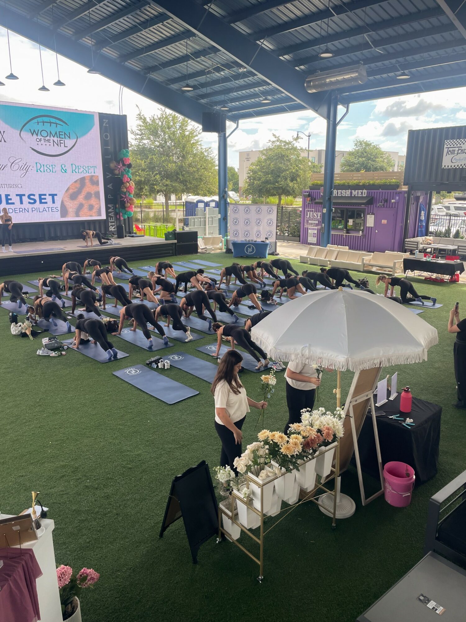 Group of people practicing yoga outdoors under a large covered area, with a stage and trees in the background.