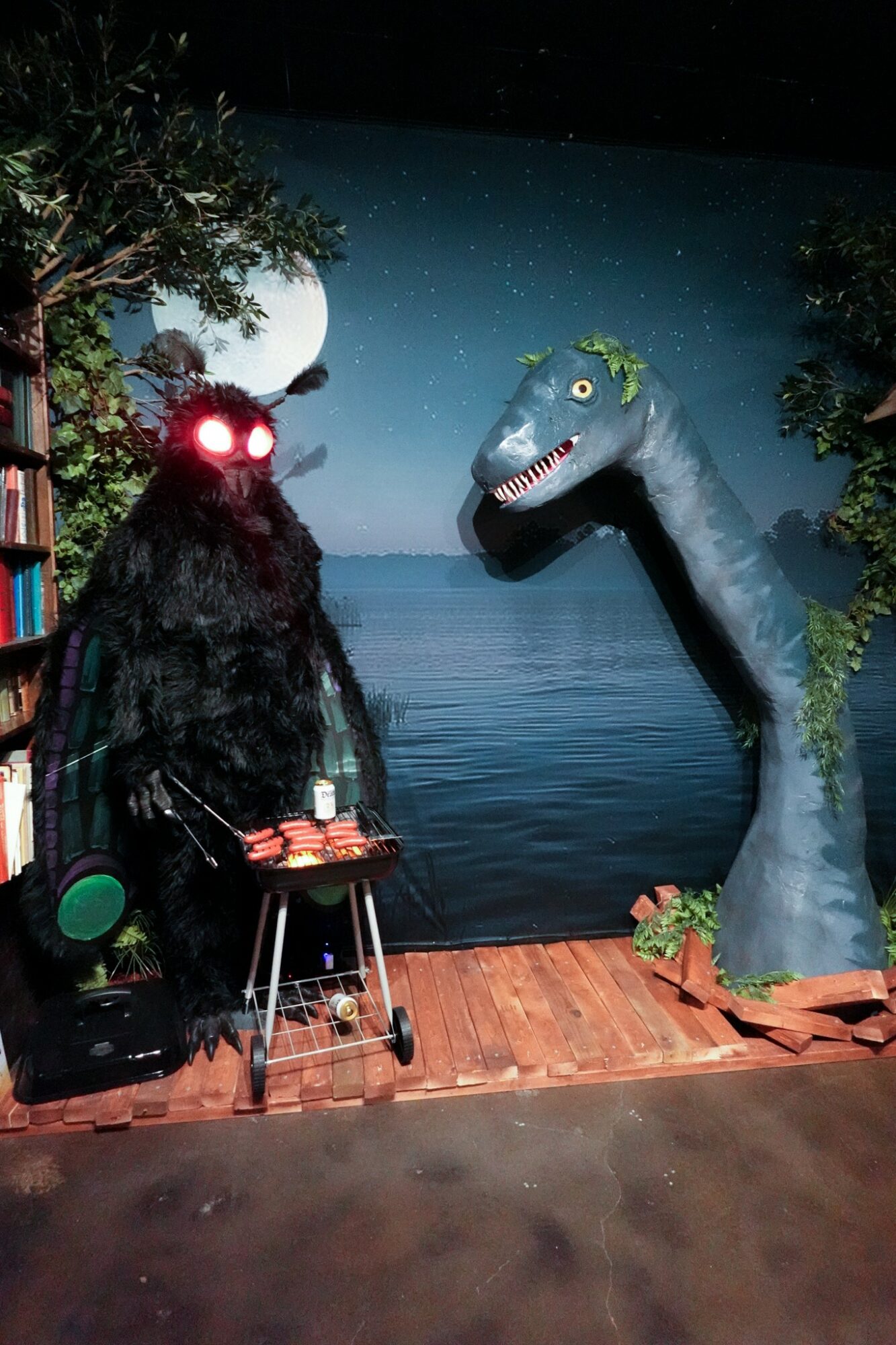 A person in a black furry costume with glowing red eyes stands next to a large dinosaur figure in front of a starry night backdrop, on a wooden floor.