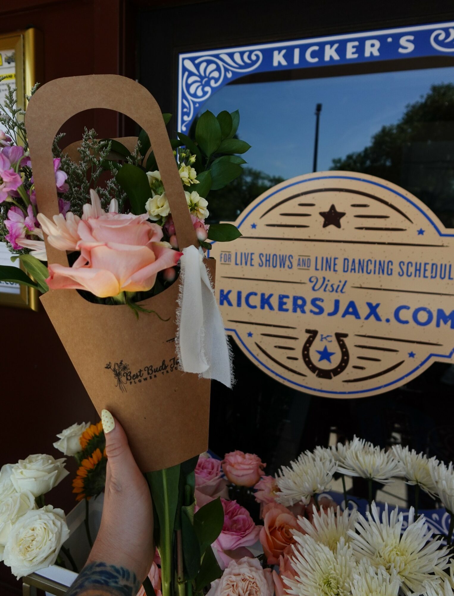 Person holds a bouquet of pink and white flowers in front of a window with a sign for Kicker's Jax, Florida. Word on sign: 'Live shows & line dancing schedule.'