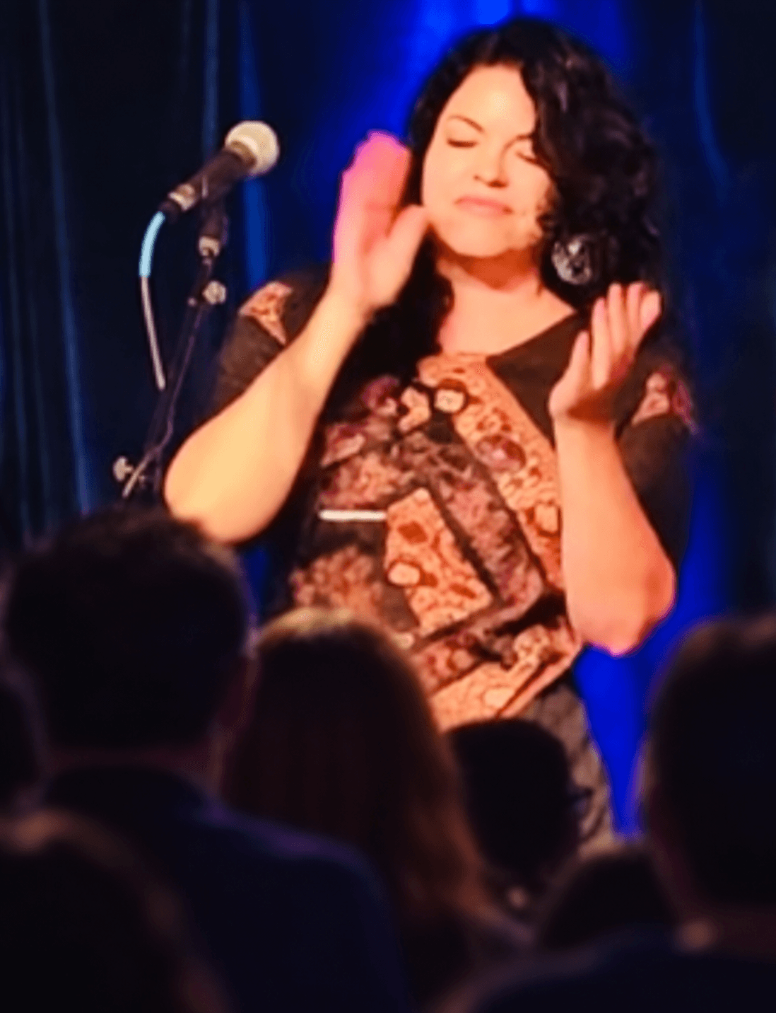 Performer with curly hair on stage, eyes closed, hands near face, microphone nearby, audience in foreground.