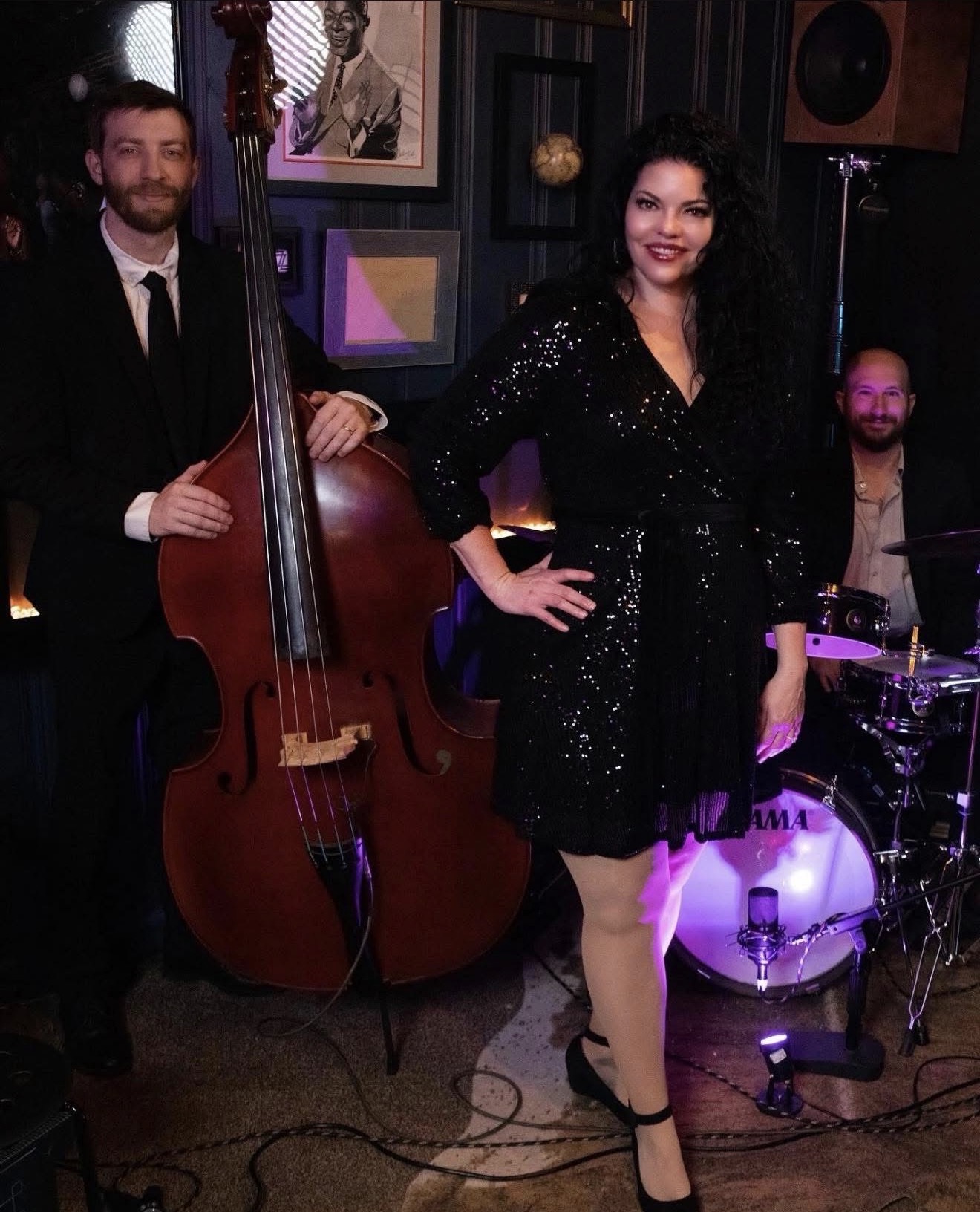 Woman in black dress with hands on hips, standing next to a double bass, smiling, with a drummer in the background.