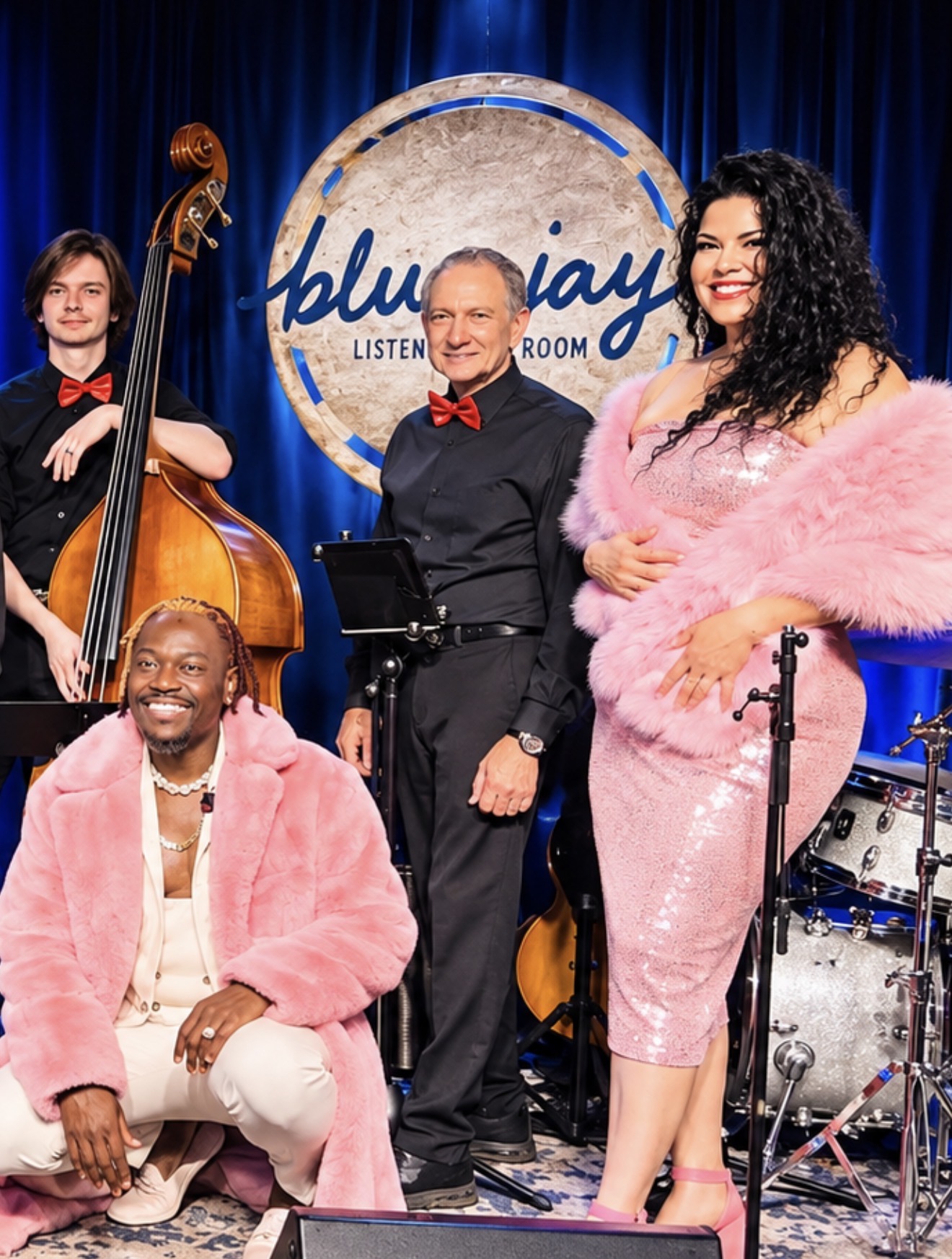 Four performers on stage, one with a double bass, two women in pink outfits, and a man in black with a red bow tie, in front of a blue curtain and a circular sign that says 'blujay'.
