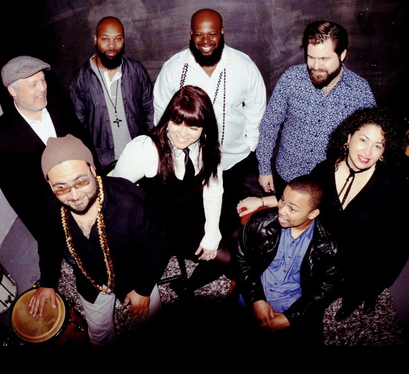 Group of nine diverse people standing together, some smiling, in a dark indoor setting.